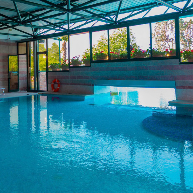 Piscina coperta con vetrate ampie, vista sul verde, e zona di passaggio verso l'esterno.