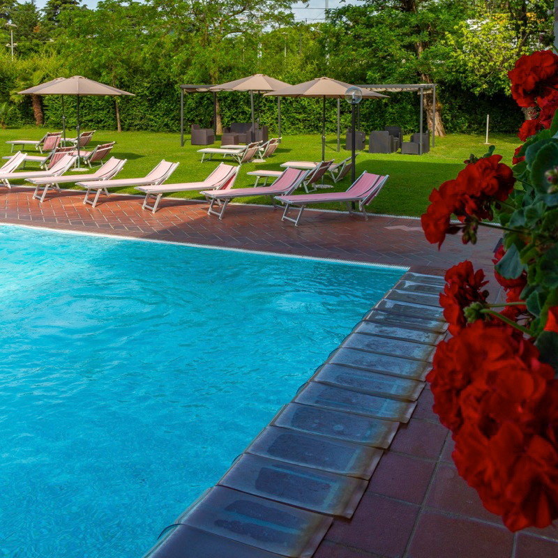 Piscina all'aperto con lettini rosa, ombrelloni beige, giardino verde e gerani rossi in primo piano.